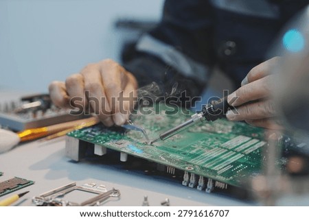 A close-up of a technician's hands using a soldering iron and tweezers to repair a green computer motherboard, with visible smoke gently rising from the hot solder point.