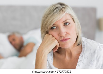 Close-up Of A Tensed Mature Woman Sitting In Bed With Man In Background At Home