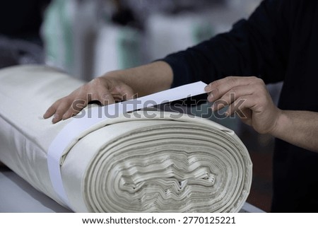 Close-up of a textile worker's hands measuring and quality controlling a large roll of white fabric in a garment factory or textile mill.