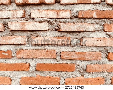 A close-up texture background of a rustic, unfinished brick wall, featuring horizontal rows of rough, reddish-orange fired clay bricks held together by thick layers of gray cement mortar.