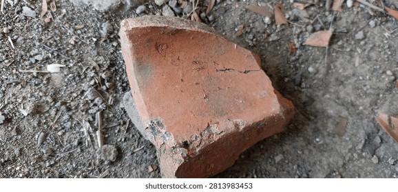 Close-up texture of a broken red clay brick lying on dry dusty soil mixed with small gravel and scattered dried brown leaves. The image features earthy tones and rough surfaces, suitable for backgrounds representing construction debris, rustic nature, or abandoned sites.