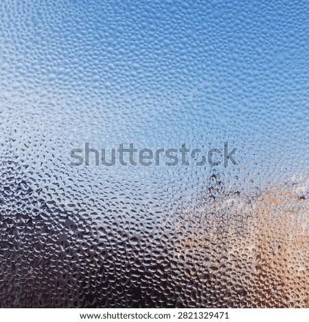 Close-up texture of countless tiny water droplets condensed on a window pane, revealing a soft, blurred background of blue sky and warm sunlight. Perfect for concepts of weather, change of season, humidity, and abstract glass textures.