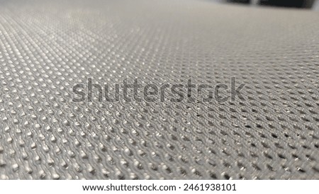 Closeup of the texture on top, silver color mesh fabric with small holes for air flow, a gray plastic sheet with many tiny round dots. The background is blurred and outoffocus. In an industrial settin