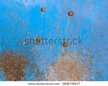 Close-up texture of weathered blue painted metal with rust stains and four corroded rivets. Peeling paint, brown corrosion patches, and rust streaks create an aged industrial background pattern.