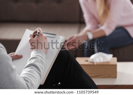 Close-up of therapist hand writing notes during a counseling session with a single woman sitting on a couch in the blurred background.