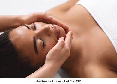 Closeup of therapist hands making facial massage for young black lady at spa salon