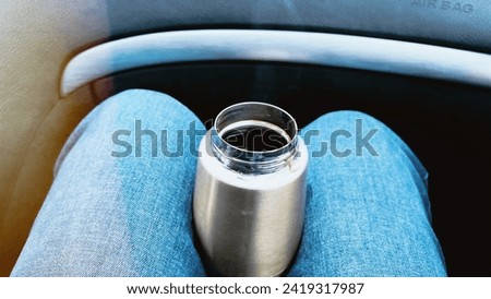 Close-up thermos between legs inside car. POV American passenger wearing jeans traveling by automobile. Auto salon. Cozy time during car ride. Soft focus. film grain pixel texture. Defocused.