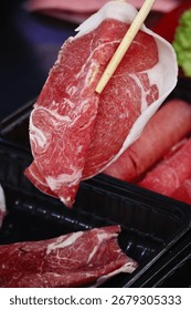 Close-up of thin lamb slice held by chopsticks above hot pot. Fresh meat ingredient for traditional Chinese hot pot cuisine.