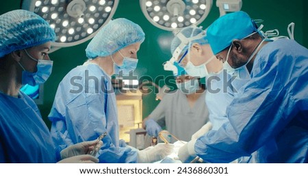 Close-up of three doctors in masks and gowns. Male doctor in binoculars. Female doctor in special cap and gloves performs operation. Medical worker holds scissors on background of operating lamp.