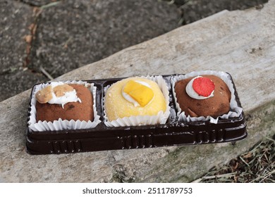 A close-up of three mochi desserts, each topped with different ingredients: biscuits, mango, and strawberry. The mochi sits in a plastic tray on a rustic wooden surface - Powered by Shutterstock