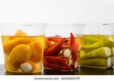Close-up Three plastic containers filled with stuffed peppers — yellow, red, and green. Each variety is submerged in oil and filled with soft cheese. The containers are placed on a black stone board