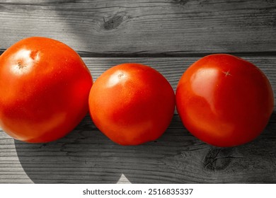 Close-up of three ripe red tomatoes arranged in a row on a rustic wooden surface illuminated by natural light. The wood texture contrasts with the smooth tomatoes. - Powered by Shutterstock