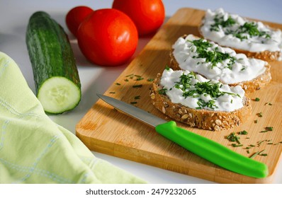 Close-up of three slices of wholemeal bread spread with butter and cottage cheese sprinkled with freshly chopped chives on a bamboo cutting board, tomatoes, cucumber and dish towel in the background - Powered by Shutterstock