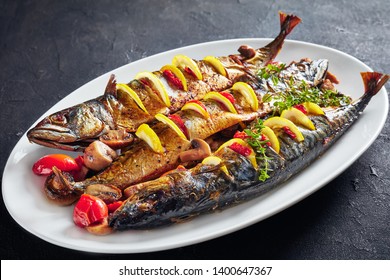 Close-up Of Three Whole Broiled Mackerels Served With Lemon, Tomatoes, Mushrooms, Spices And Herbs On A White Oval Plate, Horizontal View From Above