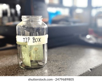 Close-up of a tip jar on a counter with some coins and a banknote inside, labeled 'Tip Box' in a café or restaurant setting.