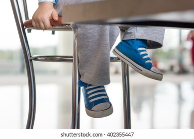 Close-up Of Toddler's Legs In Blues Sneakers Under The Table. Toddler Boy Sitting On The High Chair At The Table In The Airport. Growing Fast. Travel. Vacations.