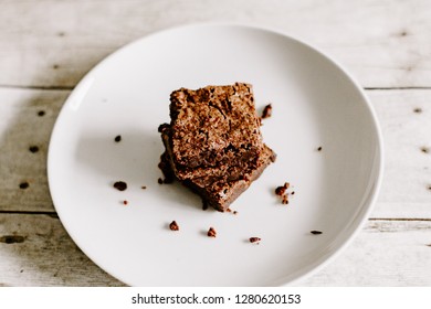 Closeup Top Down View Of A Small Stack Of Fresh Brownies Sitting On A White Plate On Top Of A Neutral Wooden Floor And Centered In Frame