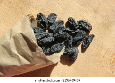 Close-up And Top View Of Black Raisins Scattered Out Of Craft Paper Wrap, The Craft Paper Wrap With Raisins Lies On The Wooden Surface, Raisins Are Illuminated By The Sun