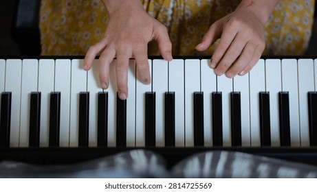 Close-up top view of a child’s hands practicing piano on a keyboard. The fingers press the white and black keys with focus and precision, while a yellow floral skirt is visible in the background. 