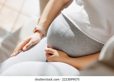 Closeup top view, pregnant woman in pajamas holds belly while sitting on gray sofa with smart watch on hand. Natural light, pregnancy concept. - Powered by Shutterstock