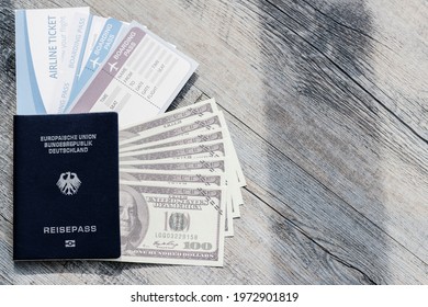 Closeup Top View Shot Of Dark Blue German Deutschland Passport One Hundred US Dollar Unfold Banknotes Cash Heap And Airline Flight Ticket Boarding Pass Arrival And Departure On Old Wood Texture Desk.