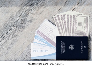 Closeup Top View Shot Of Dark Blue German Deutschland Passport One Hundred US Dollar Unfold Banknotes Cash Heap And Airline Flight Ticket Boarding Pass Arrival And Departure On Old Wood Texture Desk.