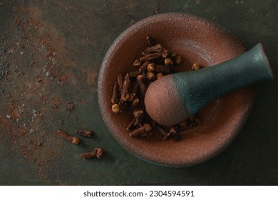 Closeup top view of a small clay mortar dish with a pestle and dried cloves, on textured rusted green background