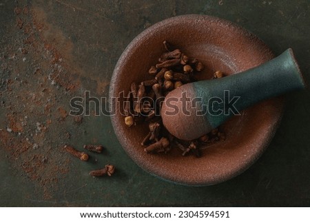 Closeup top view of a small clay mortar dish with a pestle and dried cloves, on textured rusted green background