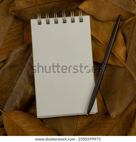 Closeup top view of a spiral blank white notepad and a black pen on brown magnolia leaves background. Square composition with copy space.