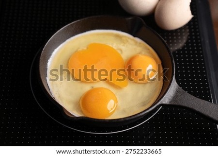 close-up top view of three raw egg yolks and whites in a small cast-iron skillet resting on a black dotted induction cooktop, warm golden hues and glossy textures with nearby eggshells, minimalist kit