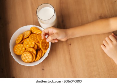Close-up top view of unrecognizable little child girl taking cookie from plate sitting at table with glass of milk. Closeup cropped shot of kid making snack during online distance learning at home.