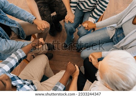 Closeup top view of unrecognizable multicultural and different ages people holding each others hands sitting in circle, support each other, during group psychotherapy session. Concept of mental health