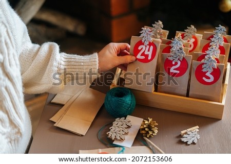 Close-up top view of unrecognizable young woman sticks number on craft bag, fastens with clothespin and puts away in wooden tray. Female making paper bags from kraft paper for advent calendar.