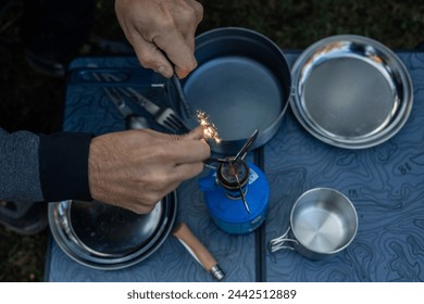 closeup A top-down perspective captures hands striking sparks from flint to ignite a camping gas stove set atop a camping table with all the necessary utensils for dinner preparation