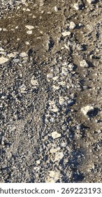 Close-up, top-down view of a gritty, textured ground surface. A mix of wet sand, gravel, and broken stones creates a natural, earthy, and abstract background.