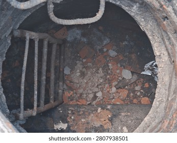 Close-up top-down view inside an open brick manhole with a rusty metal ladder and debris at the bottom.
