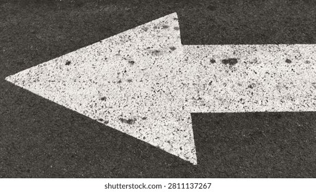 A close-up top-down view of a thick, textured white arrow pointing to the left, painted on a dark, rough asphalt road surface