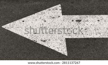 A close-up top-down view of a thick, textured white arrow pointing to the left, painted on a dark, rough asphalt road surface
