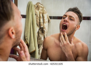 Close-up Of Topless Young Man Looking In Mirror At His Mouth And Teeth In Bathroom, Checking Their Health State