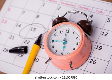 Close-up top-view photo of calendar with a datum circled with a black marker lying on it, with selective focus on an alarm clock, concept of time management at work
