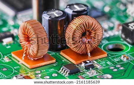 Closeup of toroidal inductors and electrolytic capacitors on electronic PCB detail with bokeh. Two ferrite core coils with copper wire winding and various surface-mount devices on green circuit board.