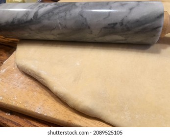 Closeup Of Traditional Rustic Kitchen Bakery Wooden Countertop, And Cutting Board With Tender Dough Rolled Out And Swirled Granite Rolling Pin. Warm Cozy Lighting Artesanal Feel, No People, With Copy 