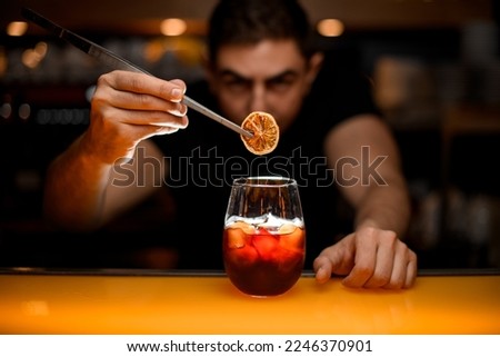 close-up of transparent glass with cold alcoholic drink and male hand accurate holding dry slice of orange with tweezers over it