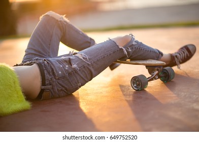 Closeup of trendy female legs relaxing on a longboard. Ripped jeans fashion. Beautiful outdoor sunlight.