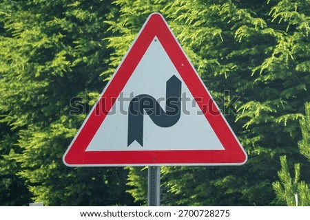Close-up of a triangular red and white traffic warning sign indicating a double bend or winding road, first to the right. The sign is mounted on a metal pole against a natural background of green pine trees. A concept image for road safety, caution, driving regulations, and navigating difficult paths or twists ahead.