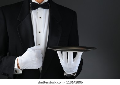 Closeup of a tuxedo wearing waiter holding a silver tray in front of his body. Horizontal format on a light to dark gray background. Man is unrecognizable.