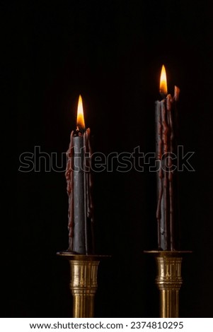Closeup of two black candles burning in golden candlestick, black background, vertical, with copy space