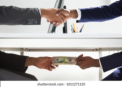 Close-up Of Two Businesspeople Shaking Hand And Taking Bribe Under Wooden Table