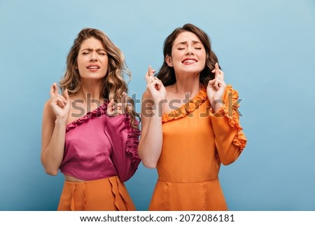Close-up of two caucasian young women crossing their fingers making wishes over isolated blue background. Blonde and brunette with closed eyes in bright clothes with bare shoulder.