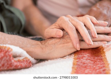 Close-up of two elderly hands gently holding each other, resting on a colorful blanket, symbolizing care and companionship.